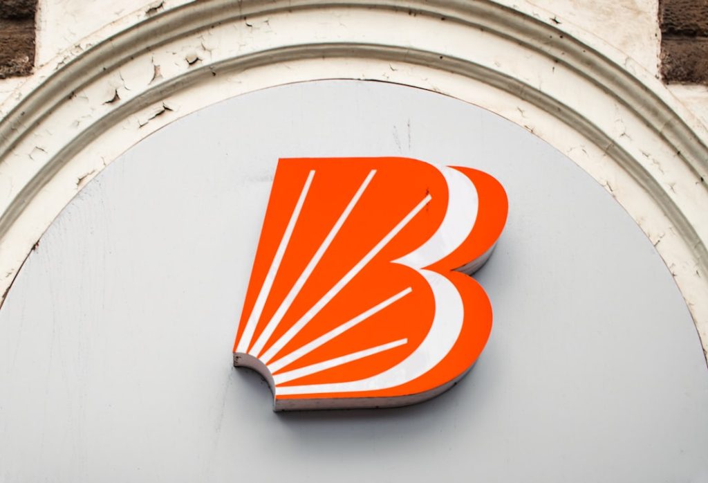 The Bank of Baroda sign stands prominently outside its branch in Ballard Estate, Mumbai—a district known for its neoclassical architecture and role as a commercial hub. As one of India’s leading public sector banks, Bank of Baroda serves a wide range of financial services, from personal banking to international trade finance. The bold orange logo and signage provide a modern corporate contrast to the historic stone buildings of the area, reflecting the coexistence of contemporary finance within Mumbai’s colonial-era infrastructure. – Foto de Zoshua Colah en Unsplash